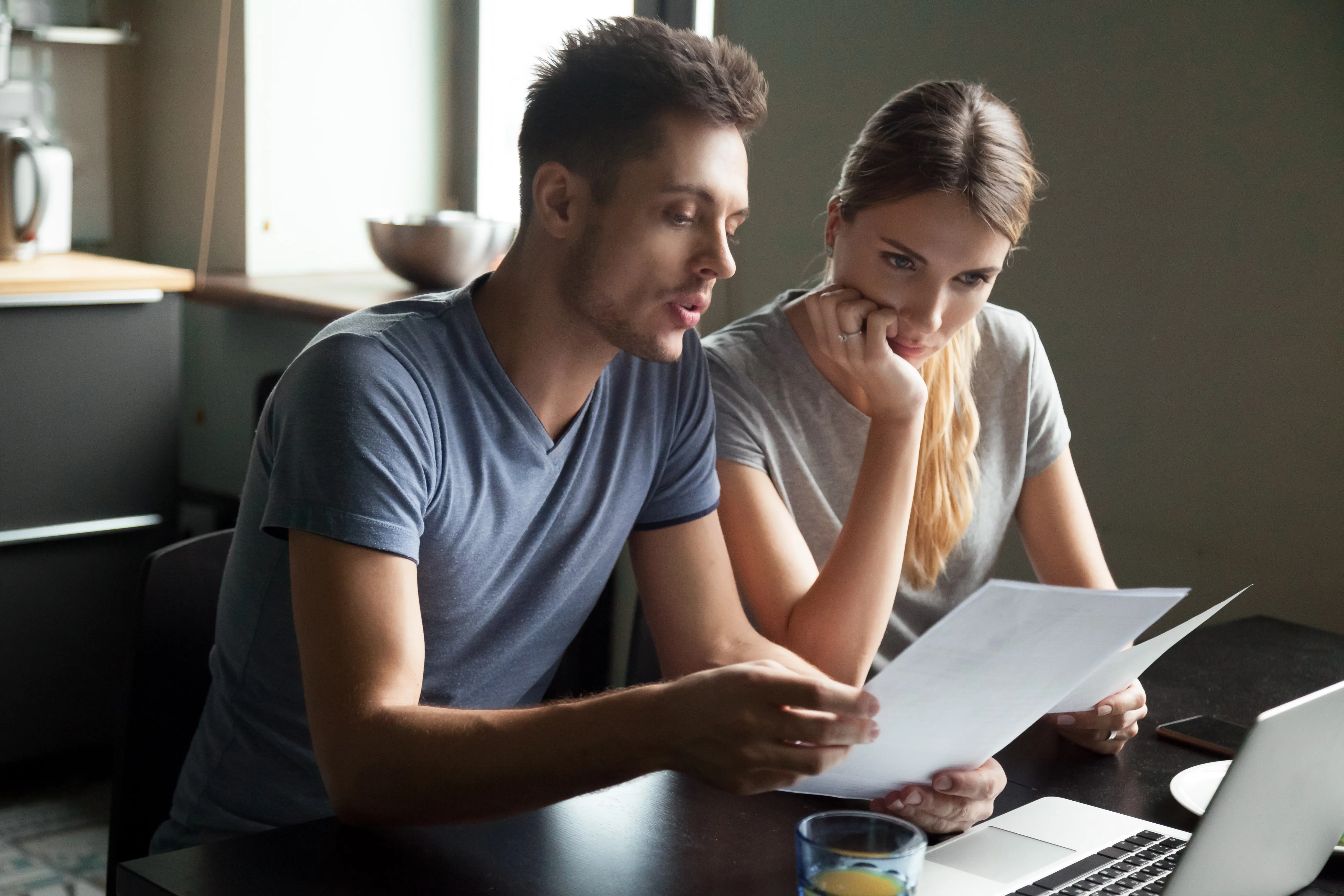 A serious, confused young couple using a laptop to review bills and bank loan documents, looking at large credit card bills, reading bad news, and confronting debt; a financial problem concept.